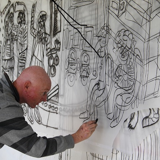 Artist John Goldsmith photographed working on a black lined artwork of people seated in an urban environment - looks like charcoal lines 